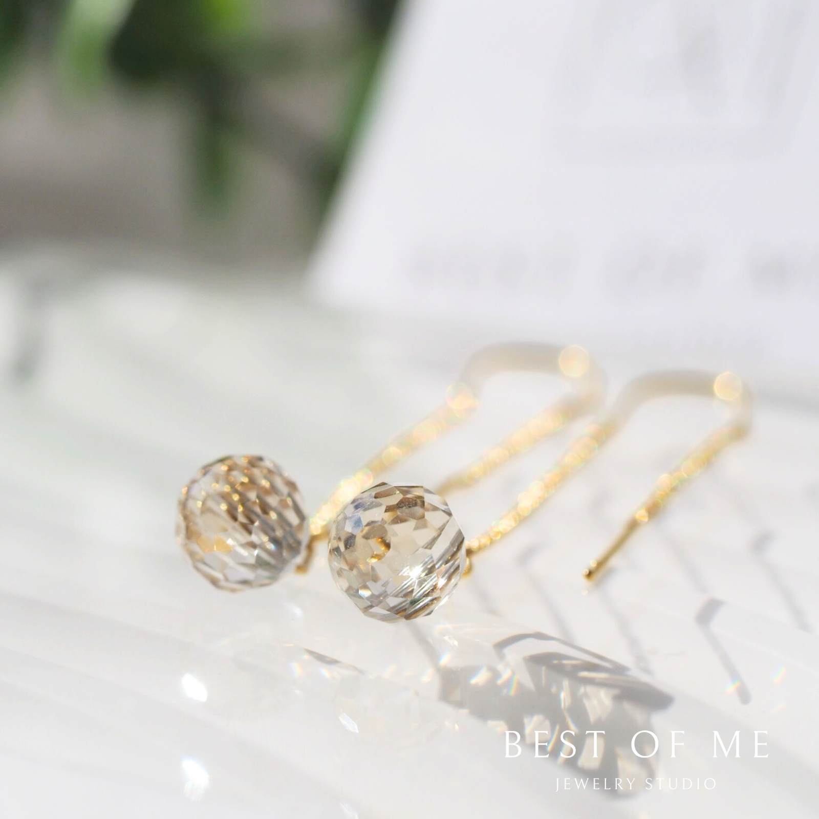 Gold earrings with light gold gemstones on a light background, featuring 'Best of Me' branding.