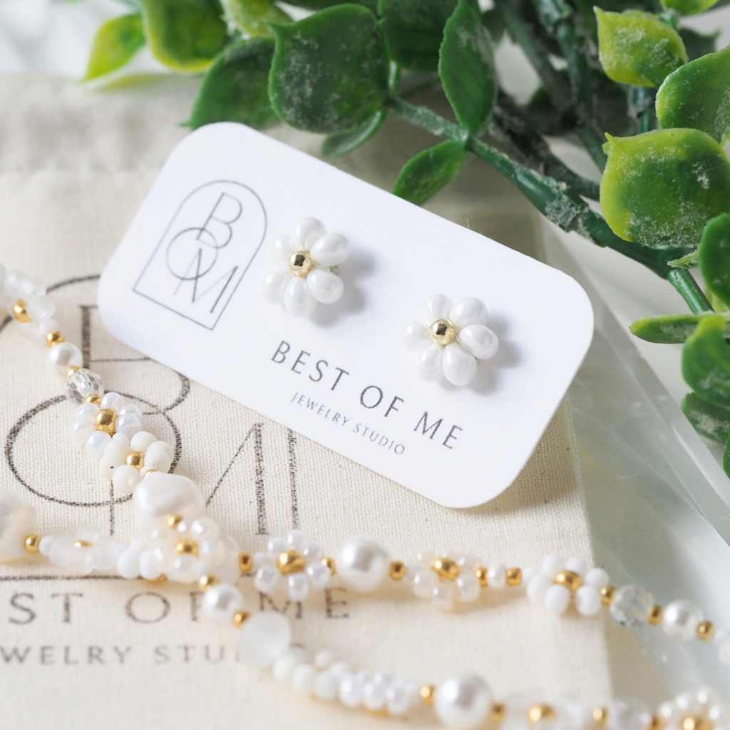 White floral earrings on a 'Best of Me Jewelry Studio' jewelry bag with greenery in the background