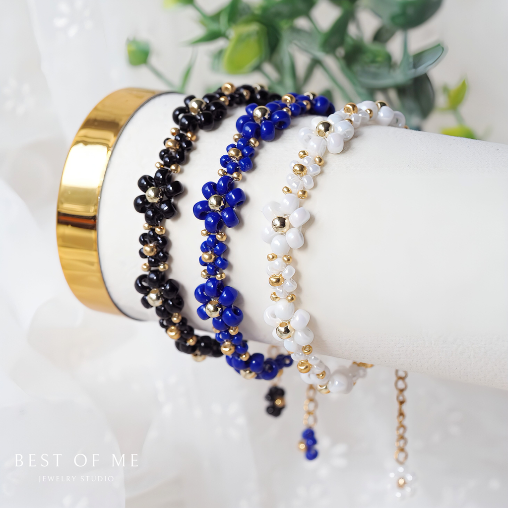 Set of beaded flower bracelets on a white mannequin hand with a gold band, against a blurred green plant background.