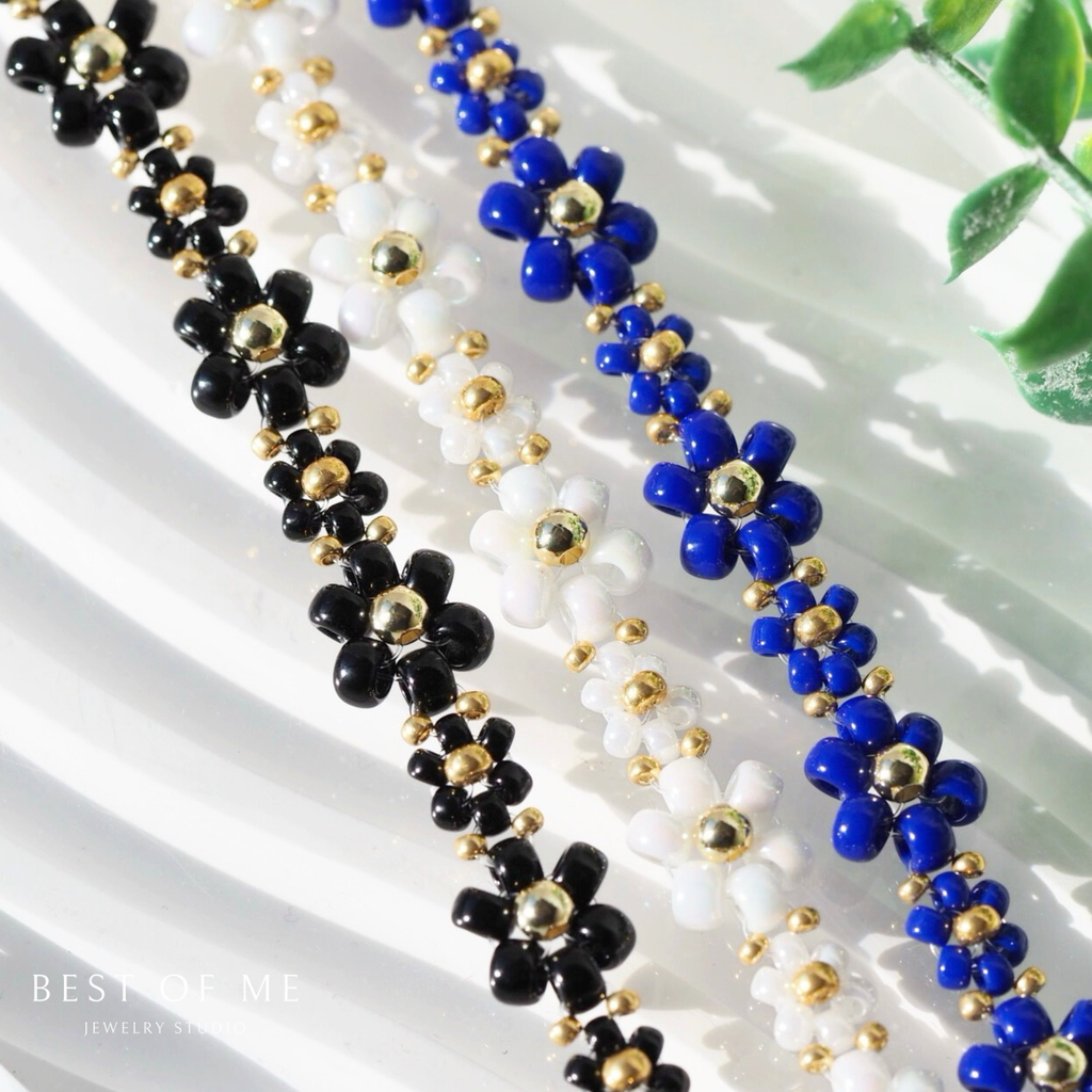 Three beaded flower bracelets in black, white, and blue with gold accents on a white surface.