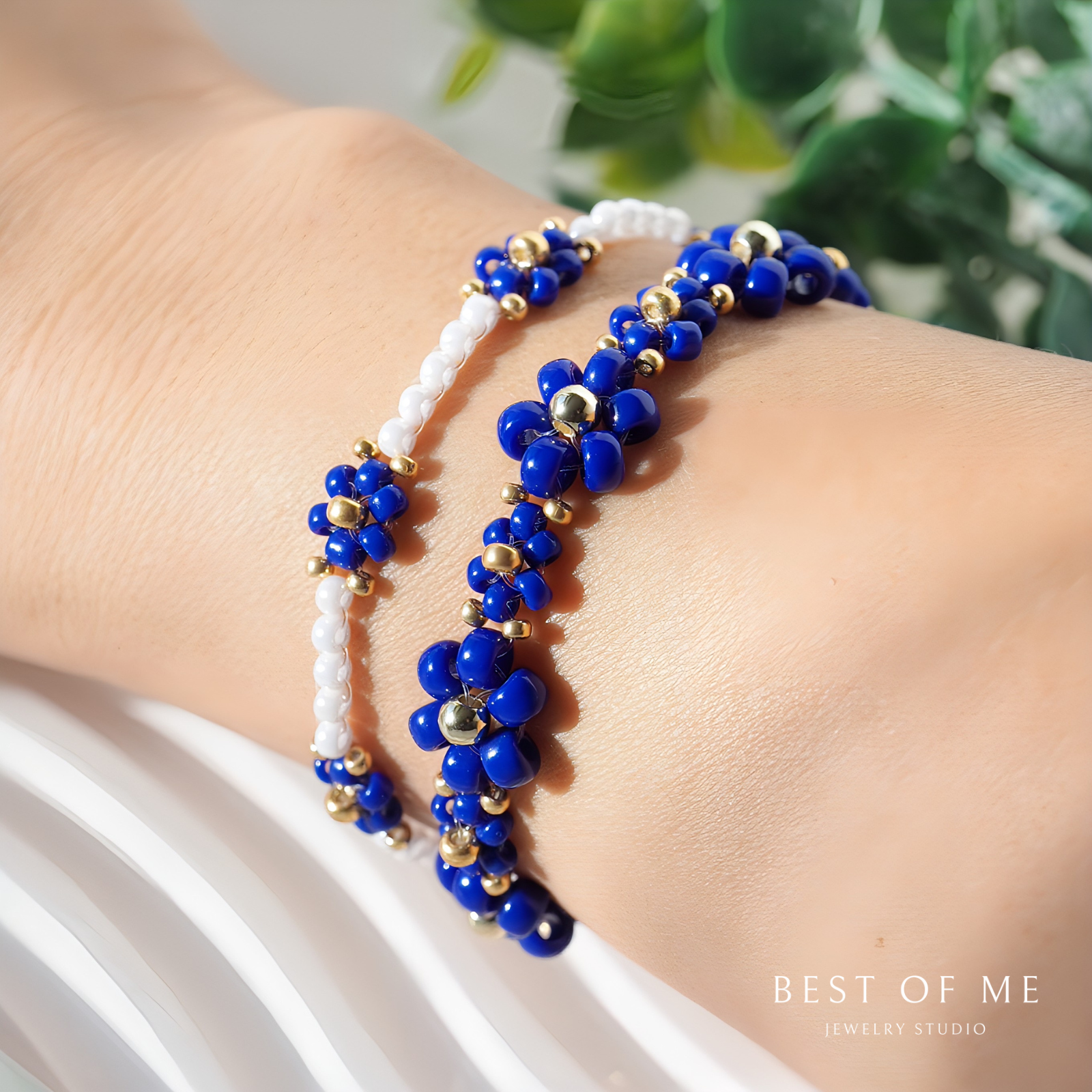 Two beaded flower bracelets, one blue and one white with gold accents, on a wrist with a blurred green plant background.