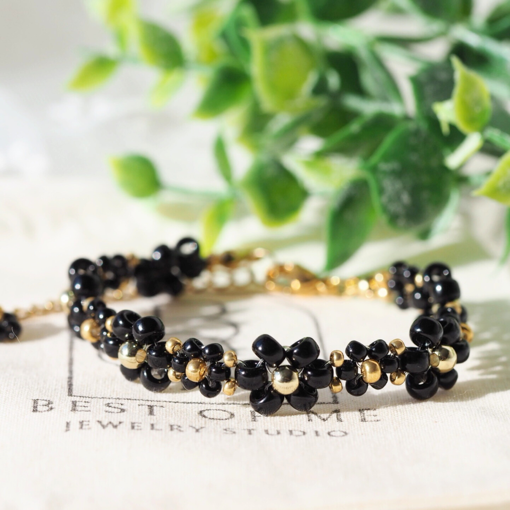 Black and gold beaded flower bracelet on a white surface with green leaves in the background