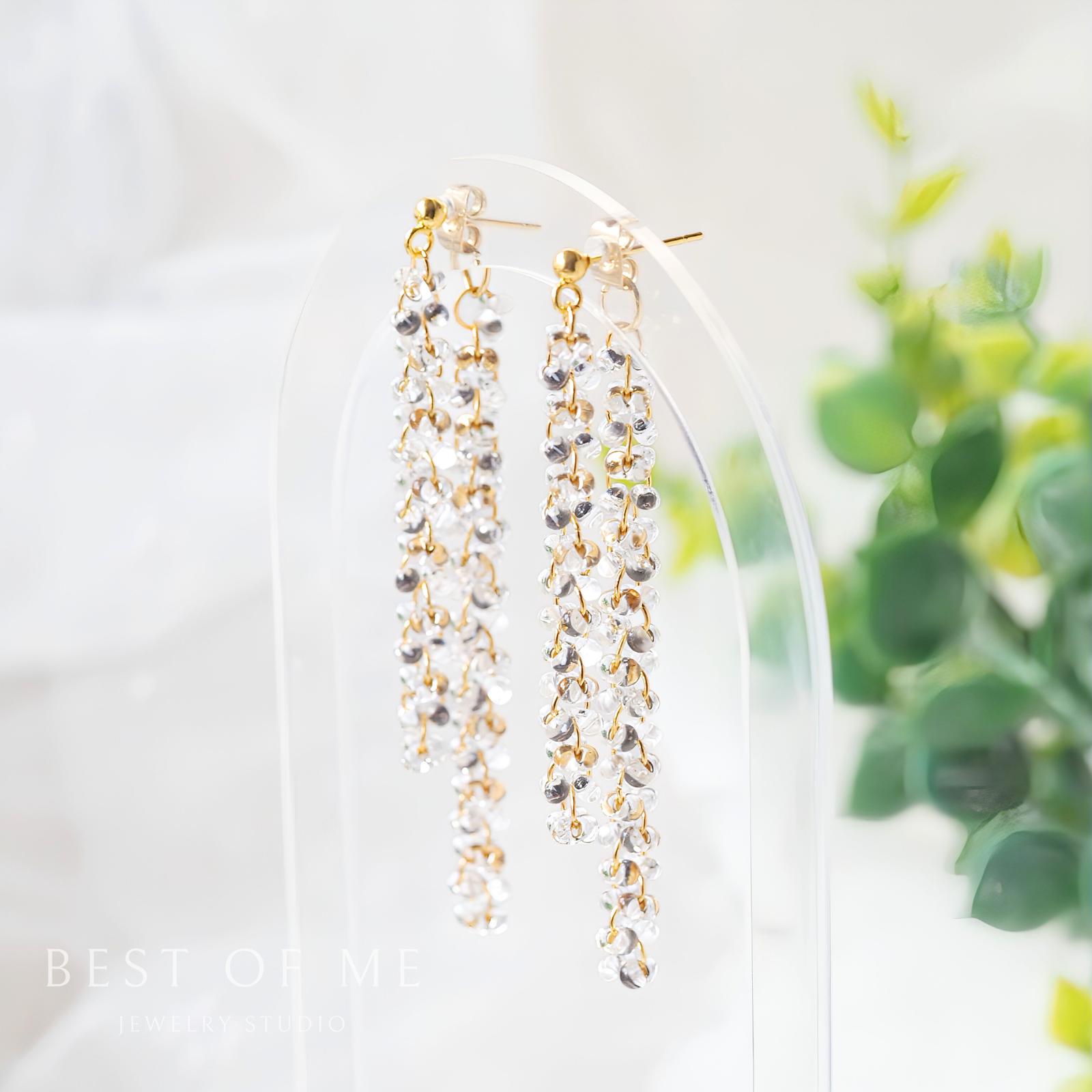 delicate beaded earrings on a clear stand with a blurred green plant background.