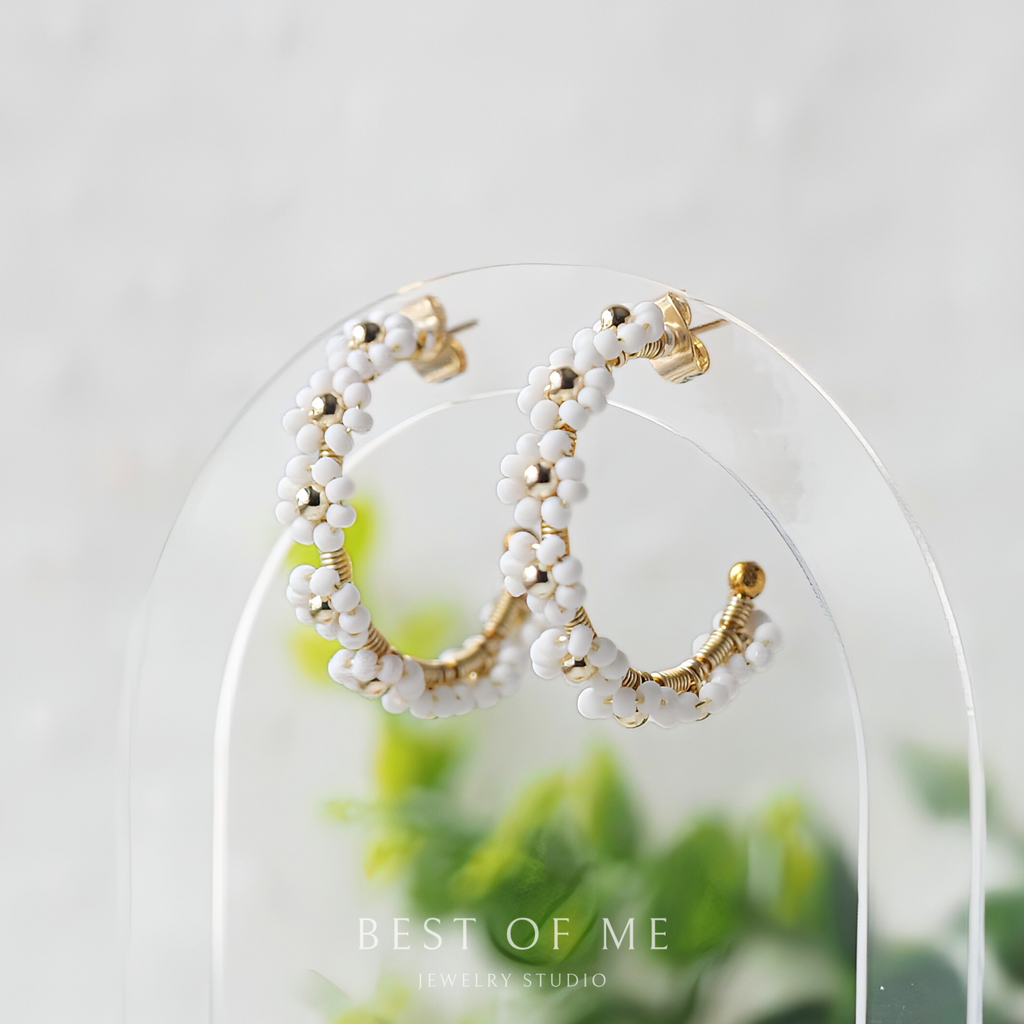 Pair of hoop earrings with white beads and gold accents on a clear stand with a blurred green plant background.