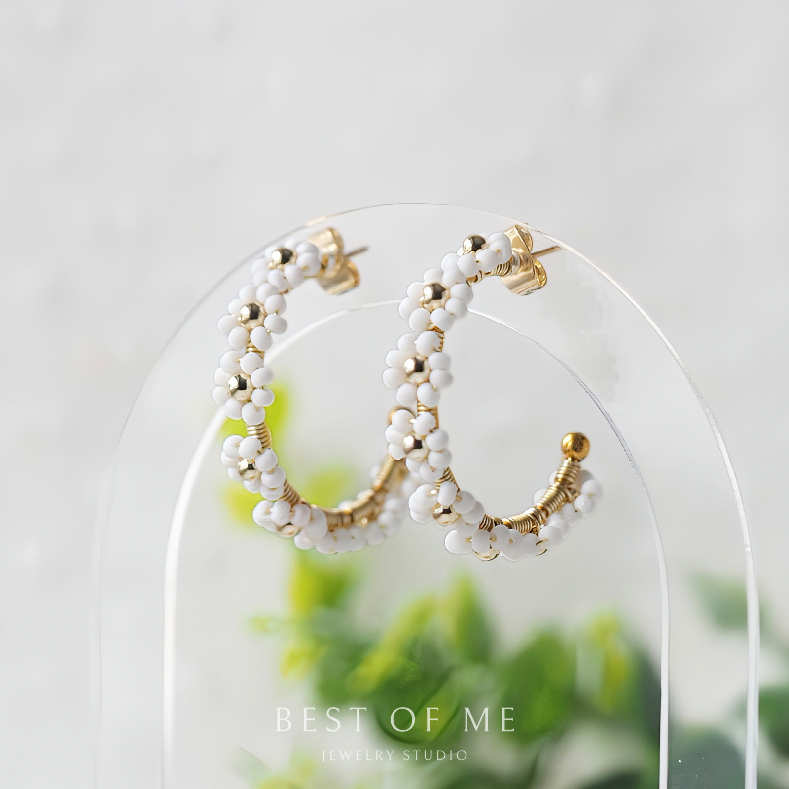 Pair of hoop earrings with white beads and gold accents on a clear stand with a blurred green plant background.