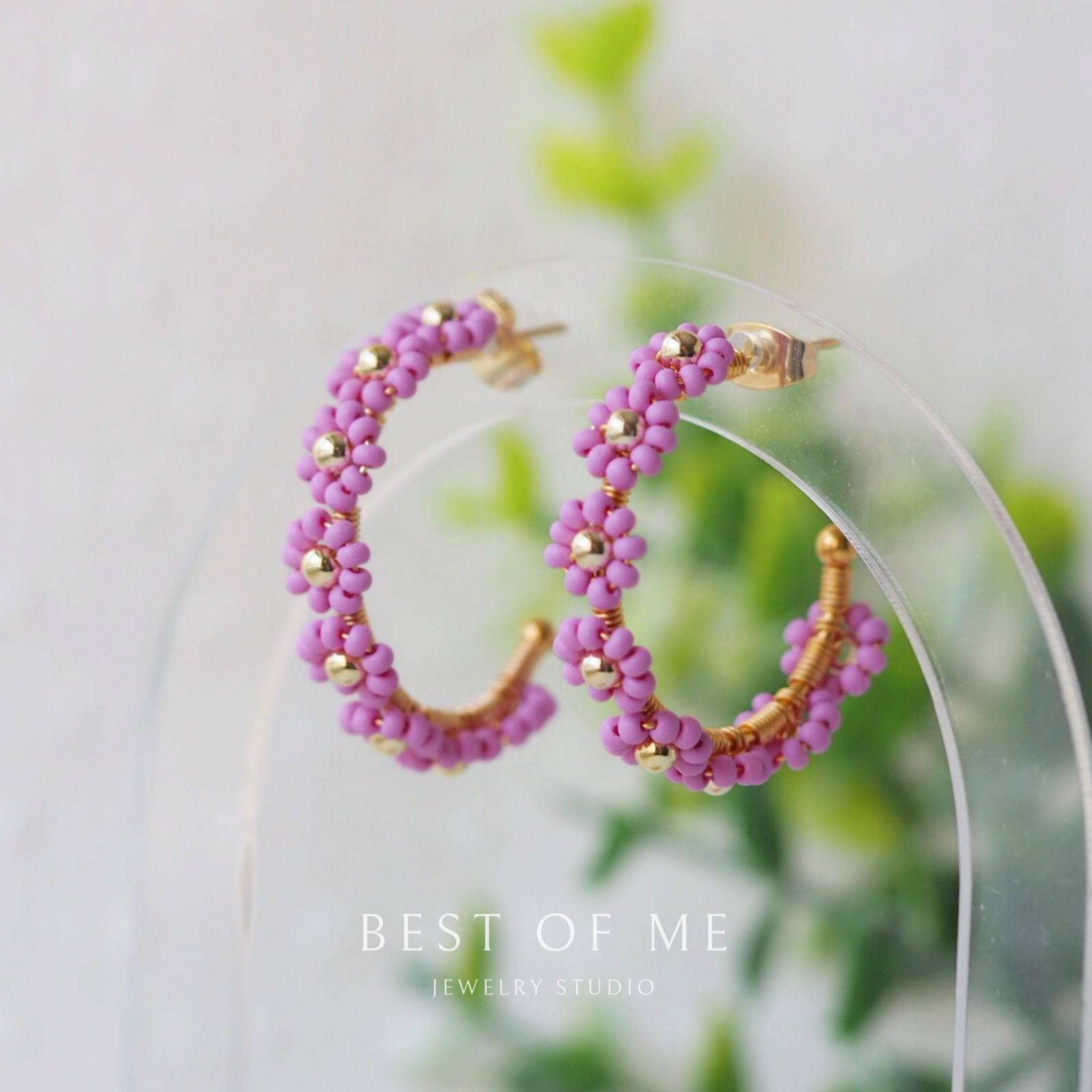 Pink beaded hoop earrings with gold accents on a clear stand, blurred green leaves in the background.