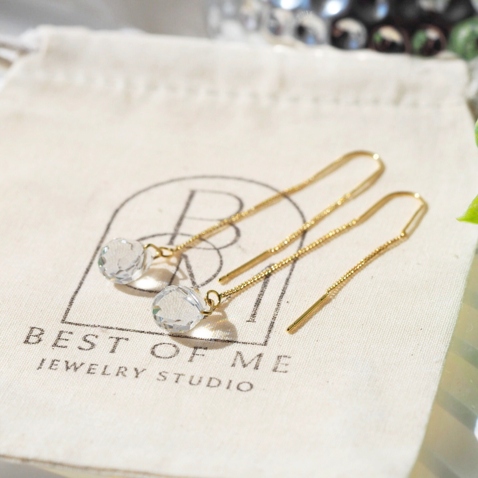 Gold earrings with clear stones on a Best of Me Jewelry Studio pouch.