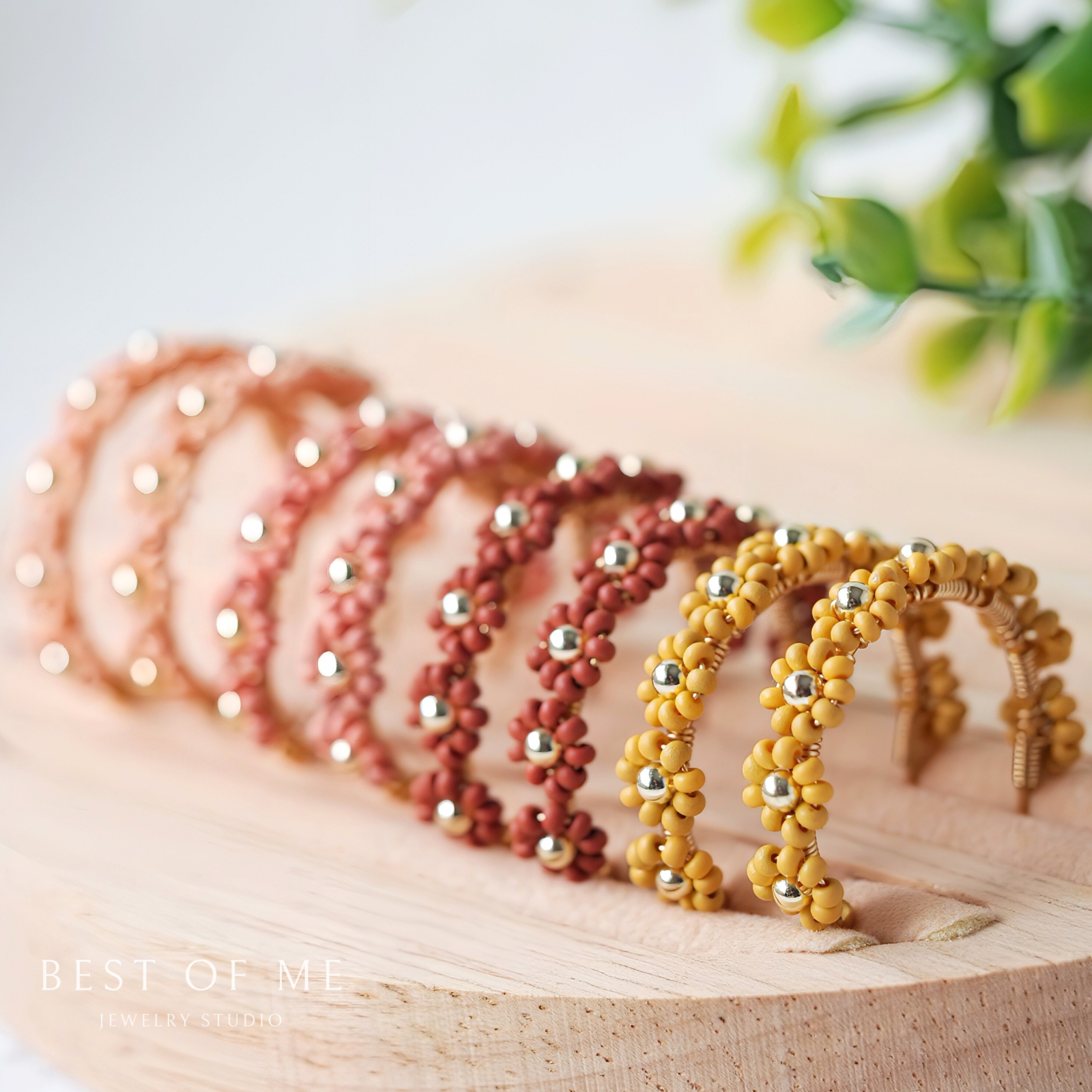 Set of beaded flower earrings in light pink, mauve, reddish brown, and mustard yellow on a wooden surface with a blurred plant in the background.
