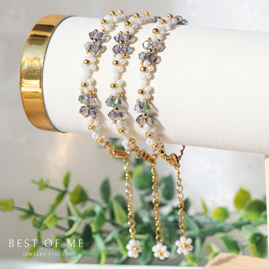 Three beaded elegant bracelets on a white cylindrical holder with a gold base, against a blurred green plant background.